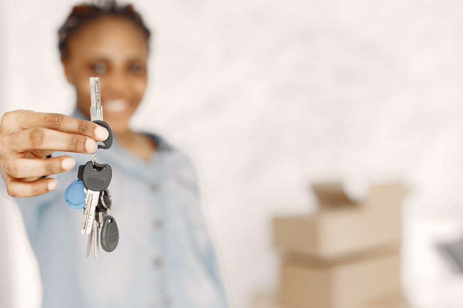 A woman with dark hair tied back, smiling and wearing a light blue shirt, holds out a set of silver keys with black plastic head, featuring a small blue key fob, towards the camera. Her fingers grip t